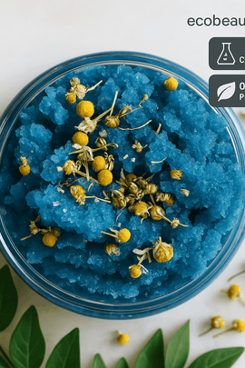 A close-up top view of a jar filled with vibrant blue sugar scrub topped with dried chamomile flowers, surrounded by green leaves and small buds, with “ecobeaut.com,” “NO CHEMICALS,” and “ORGANIC PRODUCTS” labels in the upper right corner.