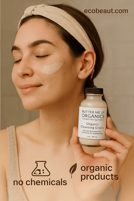 A young woman with fair skin and a beige headband gently applies a creamy organic face scrub to her cheek while holding a bottle labeled "Butter Me Up Organics – Organic Cleansing Grains." The background is a soft, neutral bathroom wall, with eco-friendly icons "no chemicals" and "organic products" at the bottom and the website "ecobeaut.com" in the top right corner.