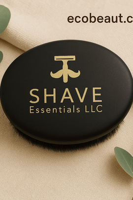 Black oval grooming brush by SHAVE Essentials LLC with gold branding on beige fabric with eucalyptus leaves — ecobeaut.com