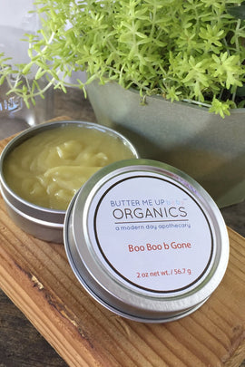 Butter Me Up Organics Boo Boo b'Gone product on a wooden board with a plant in the background