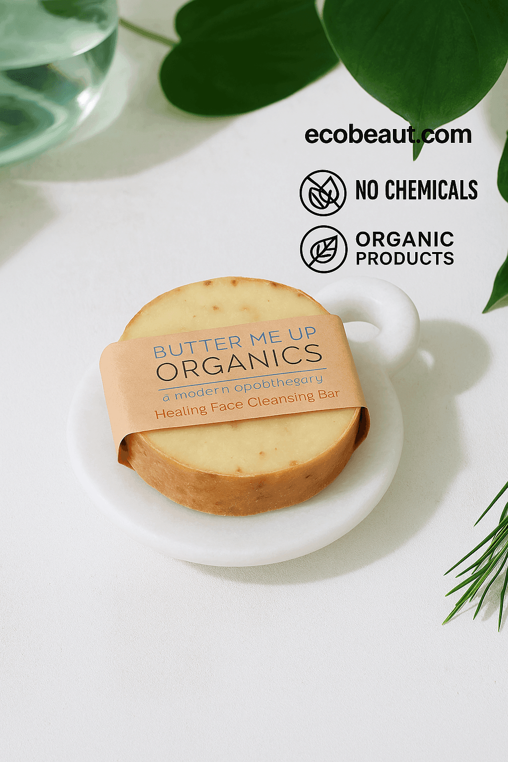 Butter Me Up Organics Healing Face Cleansing Bar on a white marble dish, surrounded by green leaves, with ecobeaut.com, “NO CHEMICALS,” and “ORGANIC PRODUCTS” icons in the top right corner.