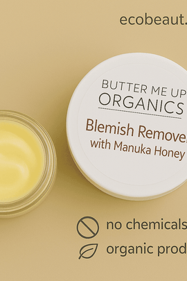 Close-up of Butter Me Up™ Organics Blemish Remover with Manuka Honey on a beige background, featuring an open jar of yellow cream, product lid with branding, and eco-friendly labels 'ecobeaut.com', 'no chemicals', and 'organic products