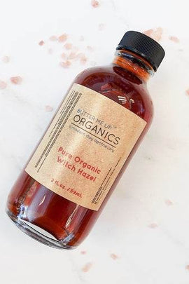 Amber glass bottle of Pure Organic Witch Hazel with kraft label on beige background, featuring ecobeaut.com in the top right and labels “no chemicals” and “organic products” below