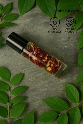"Roll-on oil bottle with dried flowers on wooden surface, surrounded by green leaves, with black eco labels and ecobeaut.com branding in the top right corner."