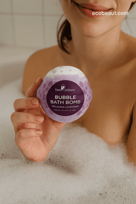 Woman holding a lavender bubble bath bomb above foamy water – natural bath product by ecobeaut.com