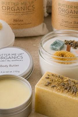 A styled product arrangement of Butter Me Up Organics bath and body items on a neutral beige surface, including a jar candle with botanicals and yellow flower, a round bath bomb, a tin of body butter, and a bar of soap.