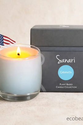 Lit candle in a glass container next to a gray box labeled 'Sanari Granita' on a beige surface.