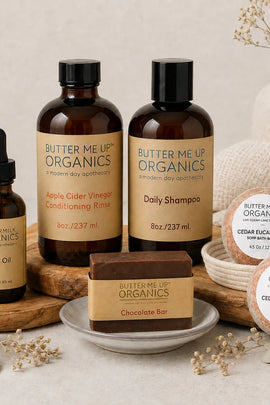 A curated product lineup from Butter Me Up Organics, including an Apple Cider Vinegar Conditioning Rinse bottle, a Daily Shampoo bottle, a Hair Oil dropper bottle, a Chocolate Bar soap bar, and two Cedar Eucalyptus soap bars in a bowl. The products are styled on a neutral beige background with decorative dried flowers, wooden boards, and a textured fabric.