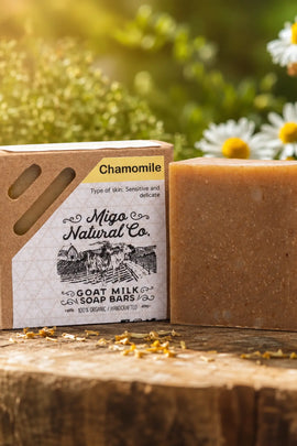 A bar of chamomile soap next to its kraft paper packaging box, styled on a rustic wooden log with daisies and green foliage in the background.