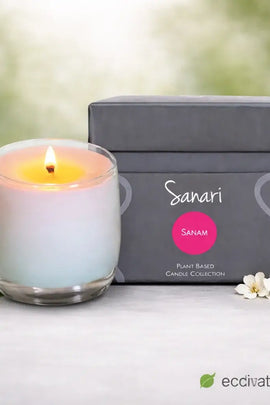 Lit candle in a glass holder next to a gray box labeled 'Sanari' on a white surface with flowers.