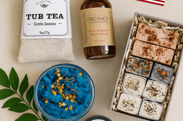 Flat lay of organic skincare products including tub tea, bath cubes, witch hazel, blue body scrub, and black drawing salve on a clean white surface, with a small American flag and ecobeaut.com logo in the corners.