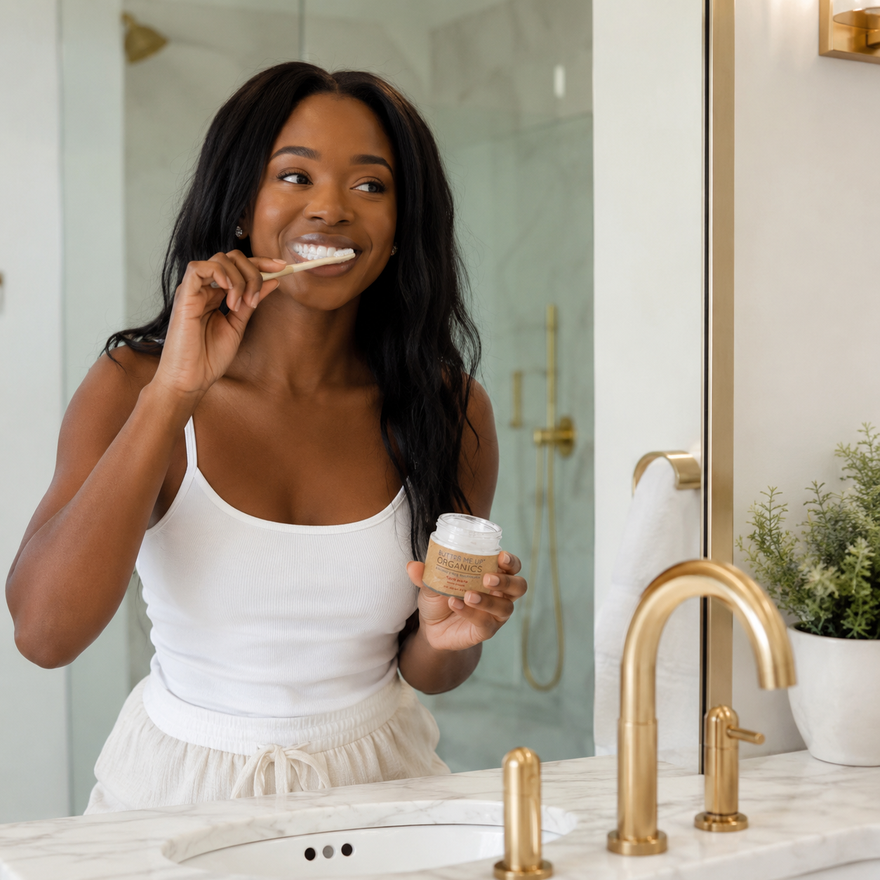 A woman with long black hair brushes her teeth while holding a small jar in a bright bathroom with gold fixtures and a white marble sink.