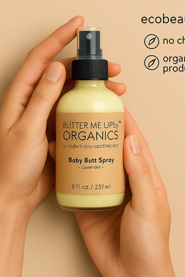 A pair of hands holds a Butter Me Up Organics Baby Butt Spray bottle with a kraft label, while the top right corner displays ecobeaut.com and eco-friendly icons for no chemicals and organic products.





