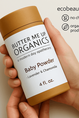 A person holds a Butter Me Up Organics Baby Powder container with a kraft texture and clean label. The top right corner shows ecobeaut.com along with icons for no chemicals and organic products.





