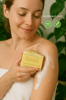 A smiling woman washes her shoulder with a Citrus Calendula soap bar by ecobeaut.com, surrounded by green plants and showing “NO CHEMICALS” and “ORGANIC PRODUCTS” icons in the upper right corner.




