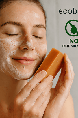 A young woman with fair skin and closed eyes smiles gently while washing her face with organic soap, producing a light foam. She holds the soap close to her cheek. In the top right corner, eco-friendly icons for "NO CHEMICALS" and "ORGANIC PRODUCTS" are displayed alongside the website “ecobeaut.com.”