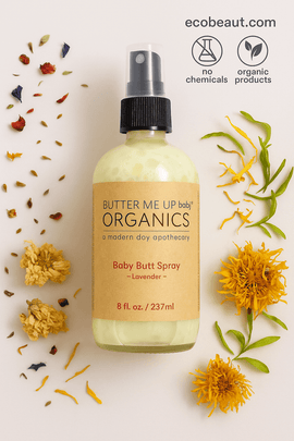 Butter Me Up Organics Baby Butt Spray bottle with kraft label lies on a light background surrounded by colorful dried flowers. Eco-friendly icons and ecobeaut.com appear in the top right corner.





