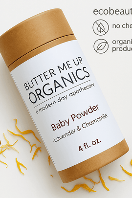 Butter Me Up Organics Baby Powder container lies on a white surface with scattered yellow petals. The top right corner features the ecobeaut.com website and eco-icons for no chemicals and organic products.




