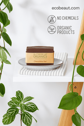  Butter Me Up Organics Chocolate Bar soap on a plate with striped cloth, surrounded by green plants, featuring “NO CHEMICALS,” “ORGANIC PRODUCTS” icons, and ecobeaut.com in the top right corner.



