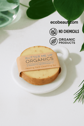 Butter Me Up Organics Healing Face Cleansing Bar on a white marble dish, surrounded by green leaves, with ecobeaut.com, “NO CHEMICALS,” and “ORGANIC PRODUCTS” icons in the top right corner.



