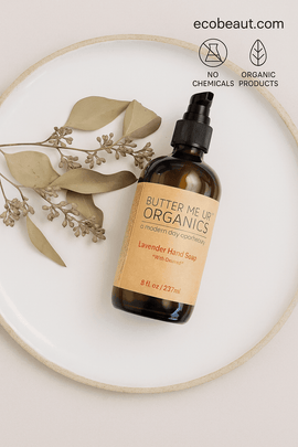 : Butter Me Up Organics Lavender Hand Soap with oatmeal placed on a ceramic plate with eucalyptus sprigs, featuring “NO CHEMICALS,” “ORGANIC PRODUCTS” icons, and ecobeaut.com in the top right corner.



