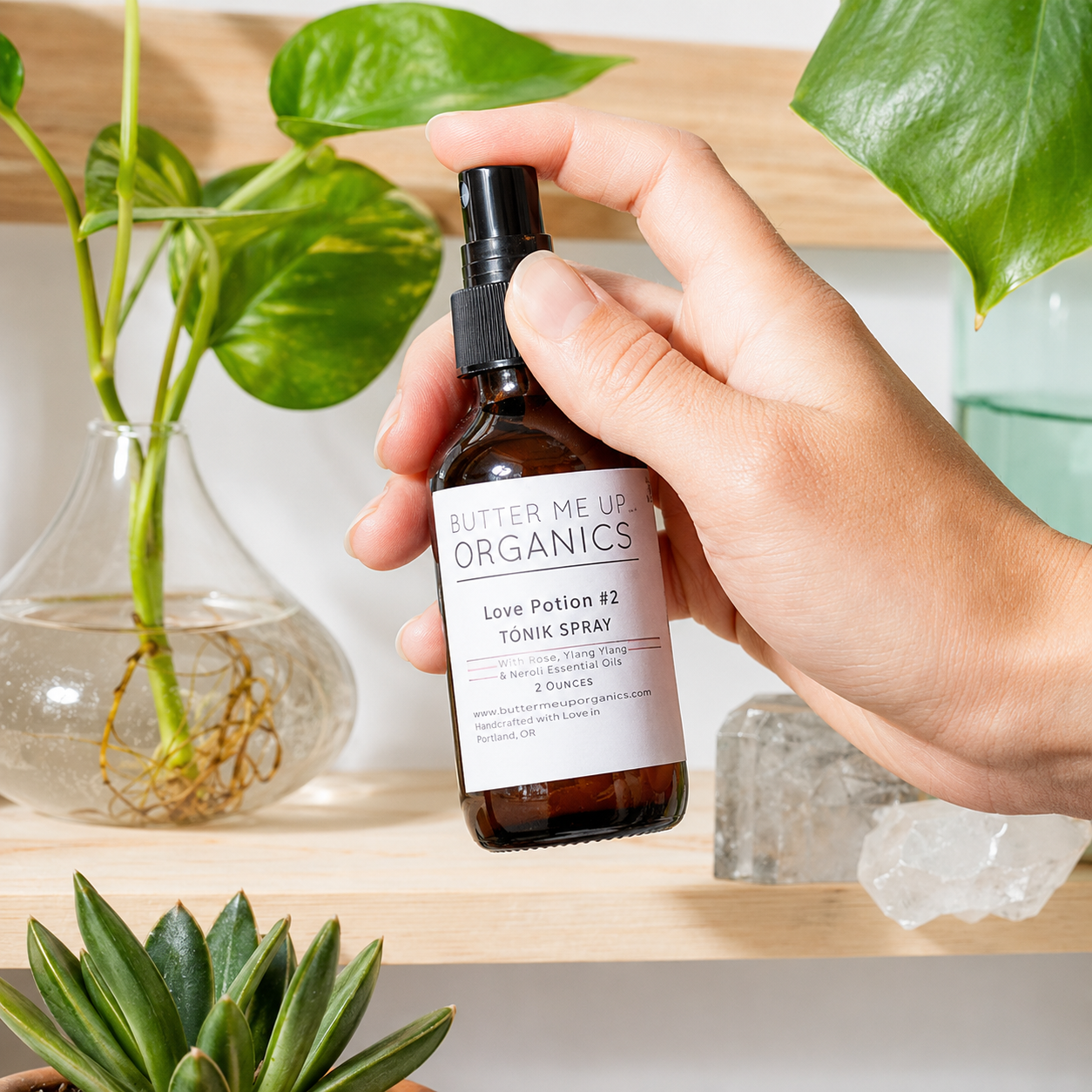A hand holds a small amber spray bottle labeled Butter Me Up Organics Love Potion #2 Tonic Spray, set against a bright, modern background with green plants and crystals.