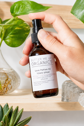 A hand holds a small amber spray bottle labeled Butter Me Up Organics Love Potion #2 Tonic Spray, set against a bright, modern background with green plants and crystals.