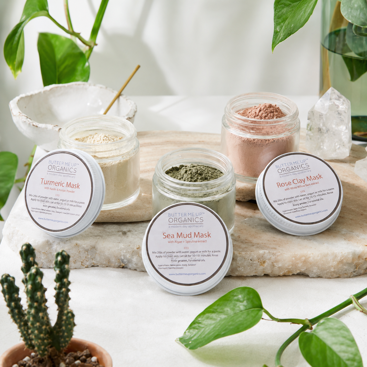 Four open glass jars of Nature's Organics face masks arranged on a stone slab with plant leaves and crystals around them. The visible products include Turmeric Mask, Sea Mud Mask, Rose Clay Mask, and a green mask, with white lids placed in front of each jar.
