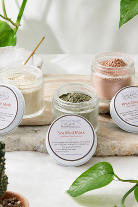 Four open glass jars of Nature's Organics face masks arranged on a stone slab with plant leaves and crystals around them. The visible products include Turmeric Mask, Sea Mud Mask, Rose Clay Mask, and a green mask, with white lids placed in front of each jar.