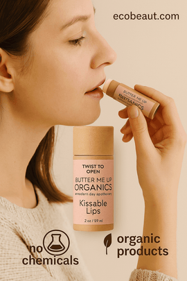Fair-skinned woman applying Butter Me Up Organics “Kissable Lips” lip balm to her lips, holding a kraft tube with a pink label. A larger version of the product is shown in the foreground. The background is soft beige, with “ecobeaut.com” in the top right corner and icons “no chemicals” and “organic products” at the bottom.