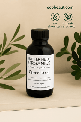 
Fair-skinned woman gently applying Butter Me Up Organics Calendula Oil to her cheek while holding the dark amber bottle, with eco-friendly icons and ecobeaut.com text in the top right corner, on a warm beige background