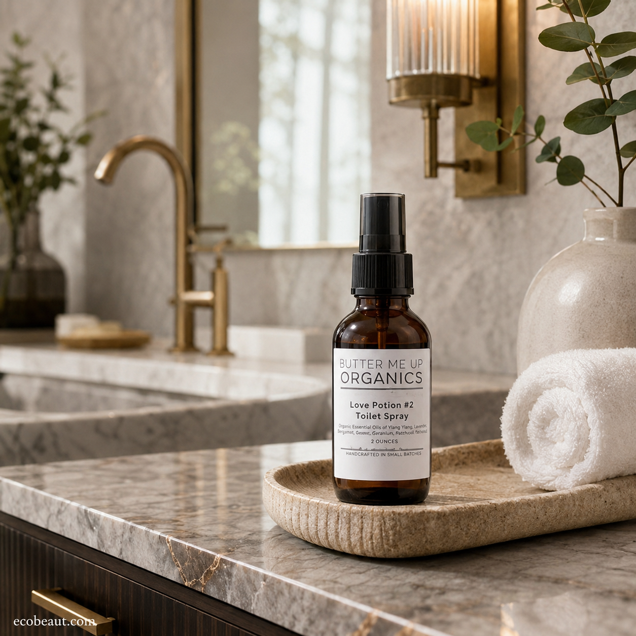 An amber glass spray bottle labeled 'Glitter Me Up Organics Love Potion #2 Toilet Spray' sits on a beige tray on a marble bathroom counter next to a rolled white towel.