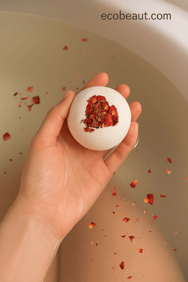 Hand holding a rose petal bath bomb above warm bath water – natural relaxation product from ecobeaut.com