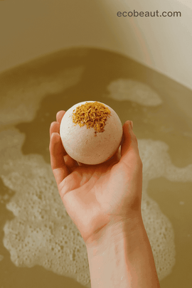 Hand holding calendula bath bomb above warm bath water, showcasing dried calendula petals on top, ecobeaut.com