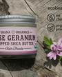 Ohana Organics Rose Geranium Whipped Shea Butter jar on a rustic wooden surface, featuring a silver lid and lavender label, surrounded by pink flowers and green leaves with ecobeaut.com, no chemicals, and organic products icons in the top right corner








