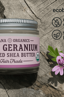 Ohana Organics Rose Geranium Whipped Shea Butter jar on a rustic wooden surface, featuring a silver lid and lavender label, surrounded by pink flowers and green leaves with ecobeaut.com, no chemicals, and organic products icons in the top right corner








