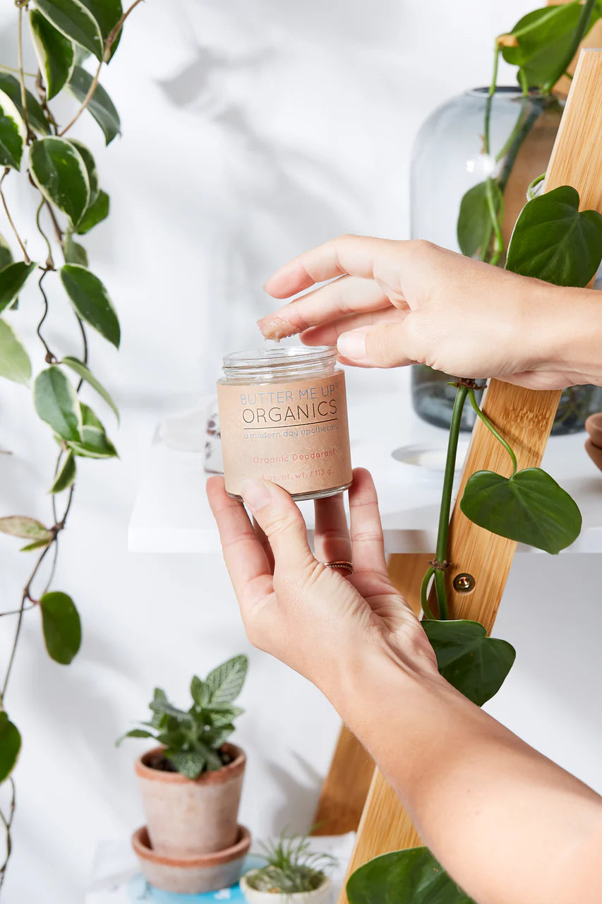A person's hands holding a glass jar candle labeled 'Blossom Organics' against a bright, modern background with green houseplants and a wooden shelf.