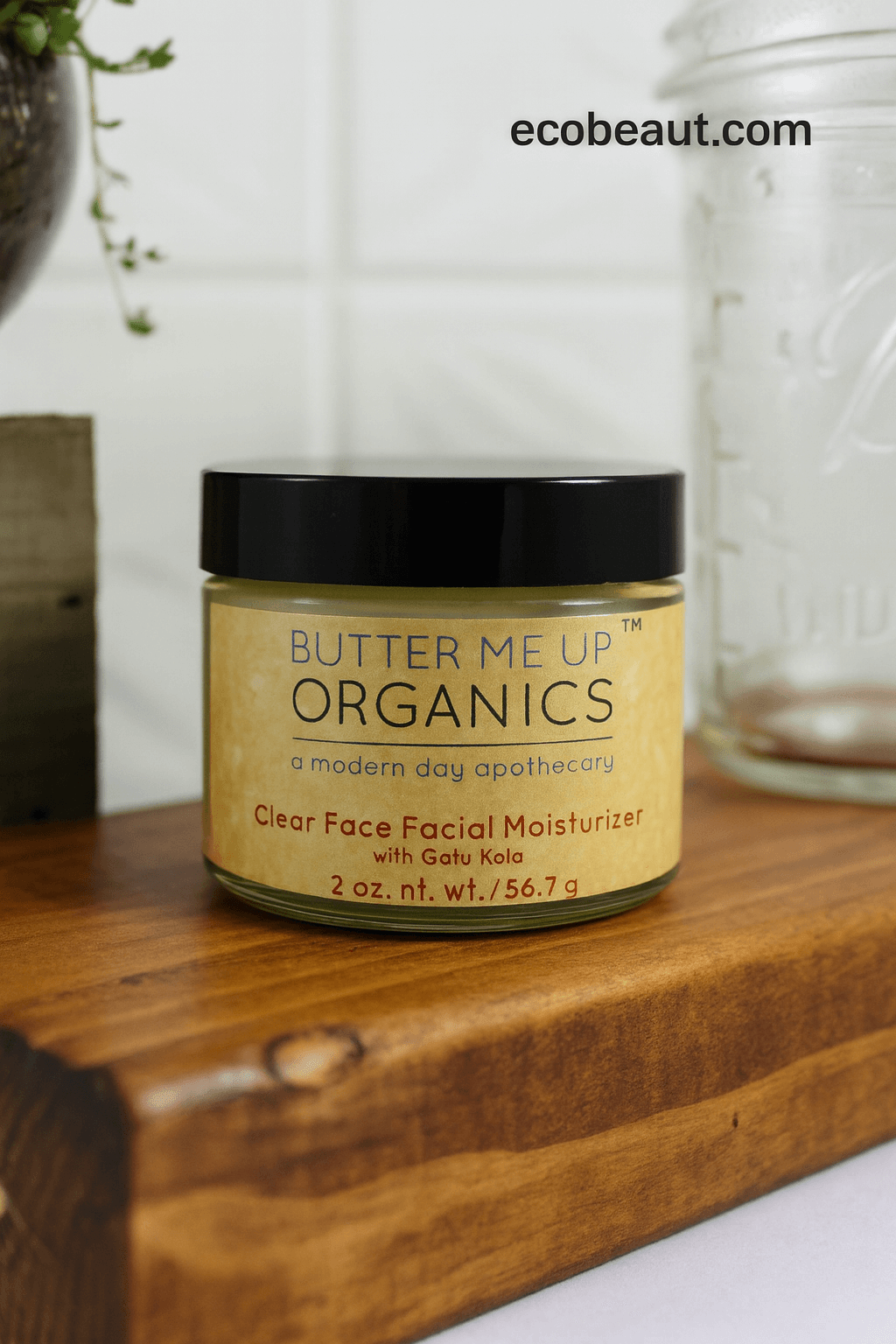 A jar of Butter Me Up Organics Clear Face Facial Moisturizer with Gaku Kola, placed on a wooden cutting board with a white tiled background and a glass jar behind it.