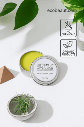 Butter Me Up Organics muscle ache salve in a tin with green leaves and a plant on a light background.