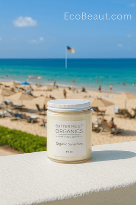 Butter Me Up Organics organic sunscreen jar on resort balcony with view of beach, pool, sunbathers, ocean waves, and distant American flag, eco-friendly sun care product branded EcoBeaut.com