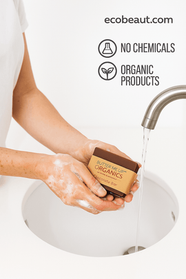 Person washing hands with Butter Me Up Organics Chocolate Bar soap under running water, featuring “NO CHEMICALS,” “ORGANIC PRODUCTS” icons, and ecobeaut.com in the top right corner.





