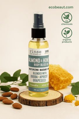 Rinse Bath & Body Co. Almond & Honey Body Bliss Oil bottle placed on a wooden slice with almonds and honeycomb, set against a warm, earthy background with eco-friendly icons and ecobeaut.com in the top right corner