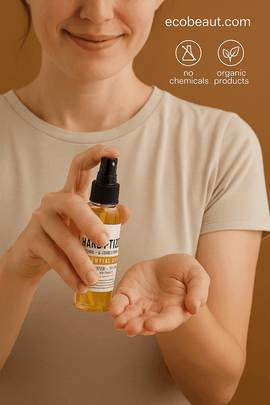 Smiling light-skinned woman spraying Handy-Tizer natural alcohol-free cleanser onto her palm, holding a small clear bottle with yellow liquid, against a warm brown background with eco-friendly icons and ecobeaut.com in the top right corner