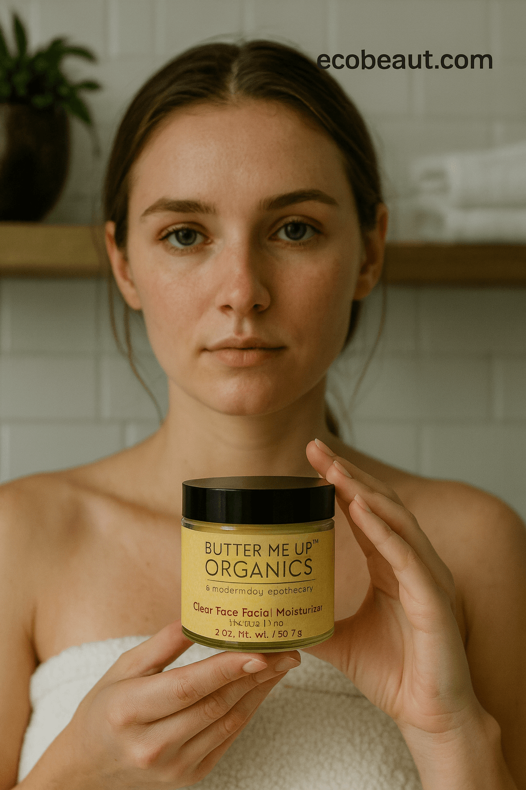 A woman wrapped in a white towel holds a jar of Butter Me Up Organics Clear Face Facial Moisturizer in front of a softly blurred bathroom background.