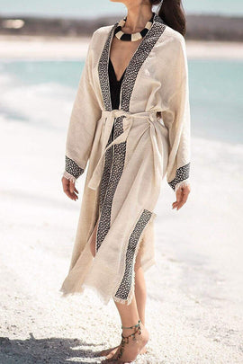 A woman standing on a beach wearing a beige linen kimono with geometric patterns, paired with a black swimsuit top and a necklace.