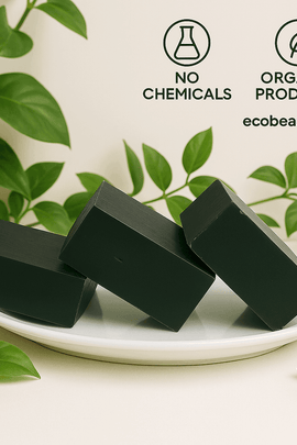 Three charcoal soap bars by ecobeaut.com arranged on a white plate surrounded by green leaves, with “NO CHEMICALS” and “ORGANIC PRODUCTS” icons and the website in the upper right corner.



