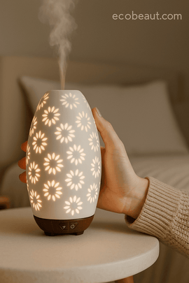 Woman placing drops of essential oil into daisy ceramic diffuser on table, with soft lighting and ecobeaut.com branding in the corner