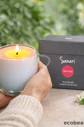 Hand holding a lit candle next to a Sanari product box with a blurred green background