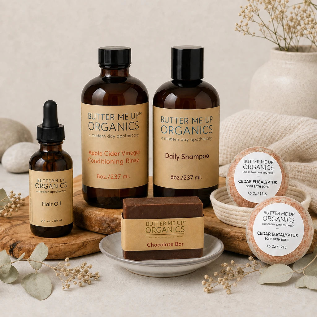 A curated product lineup from Butter Me Up Organics, including an Apple Cider Vinegar Conditioning Rinse bottle, a Daily Shampoo bottle, a Hair Oil dropper bottle, a Chocolate Bar soap bar, and two Cedar Eucalyptus soap bars in a bowl. The products are styled on a neutral beige background with decorative dried flowers, wooden boards, and a textured fabric.