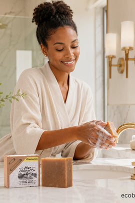 A woman in a cream bathrobe washes her hands at a marble bathroom sink, with a bar of soap and its packaging box in the foreground. The box is labeled 'Charmeroi' and 'Natural Co.